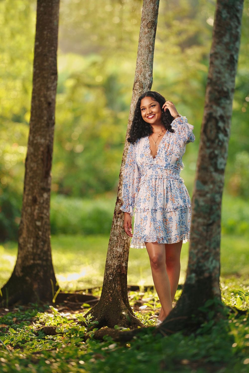 A person in a floral dress poses playfully among trees in a sunlit forest setting.