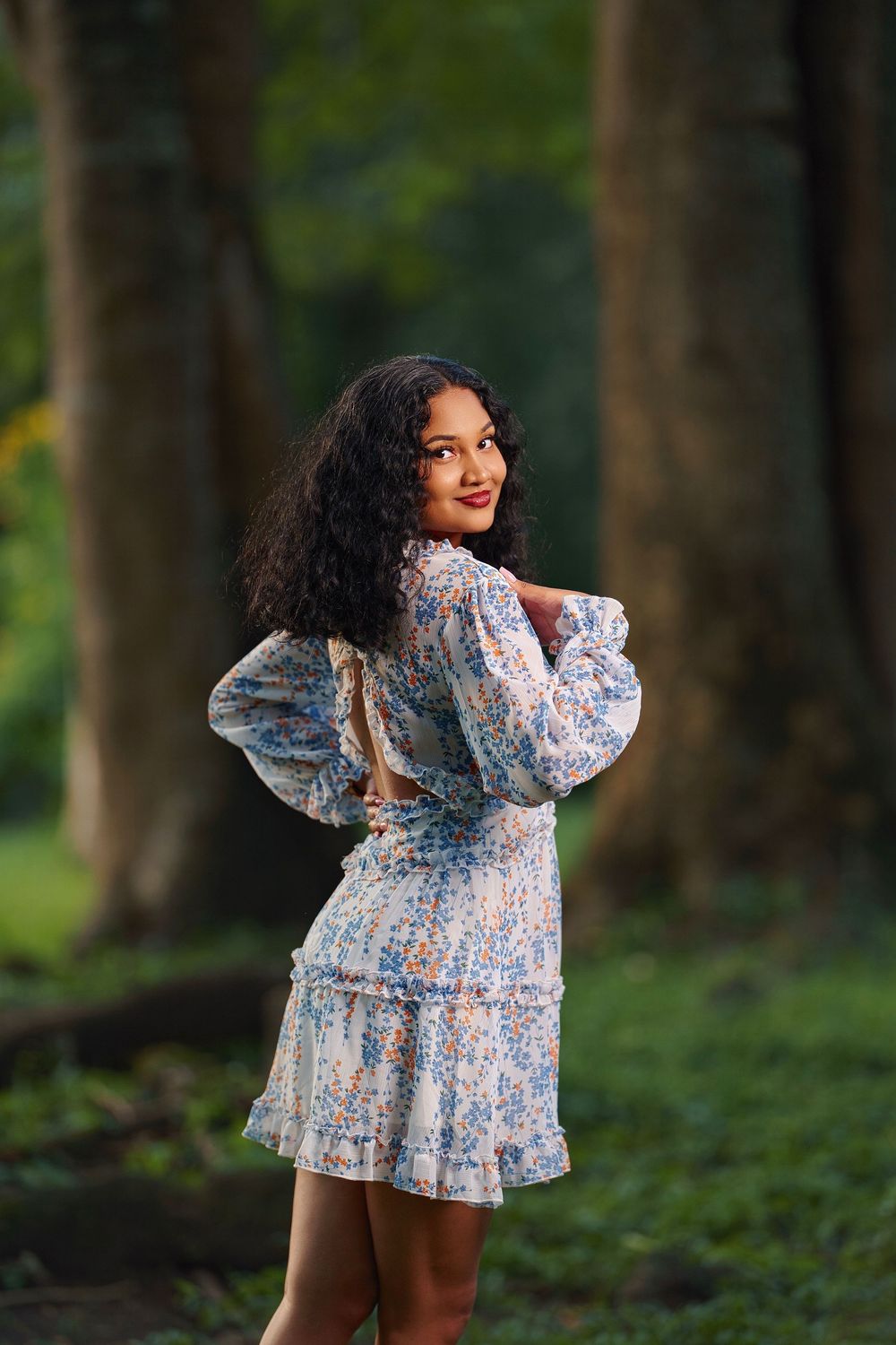 A person in a floral print mini dress poses playfully in a wooded outdoor setting with green foliage in the background.