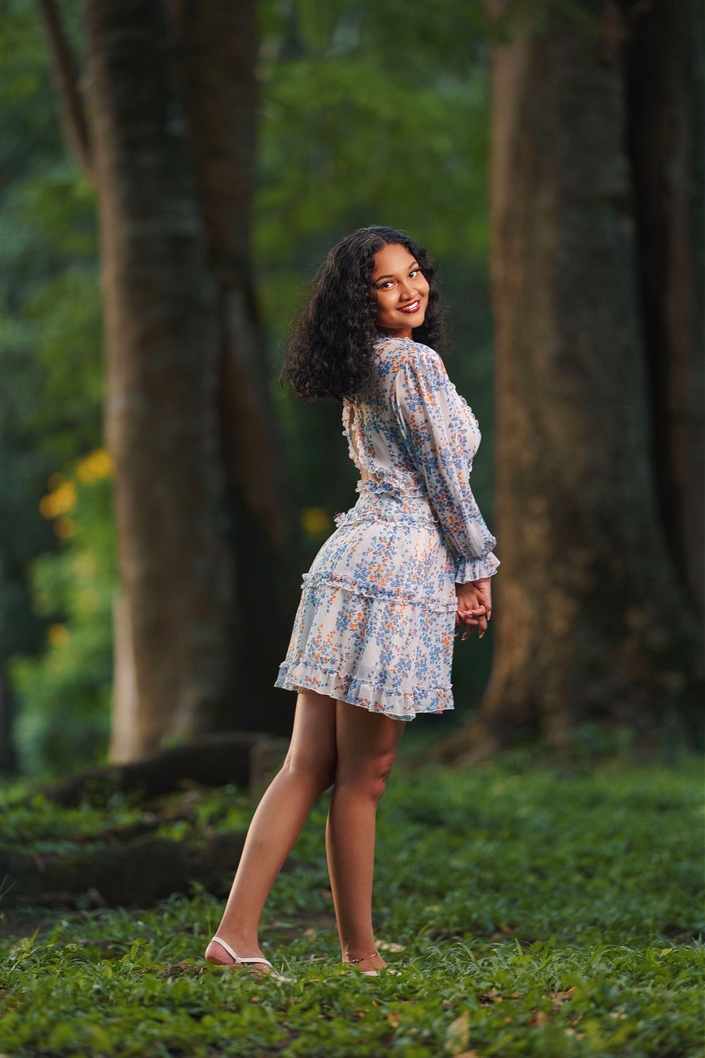 A person in a floral dress poses playfully in a lush forest setting with green foliage in the background.
