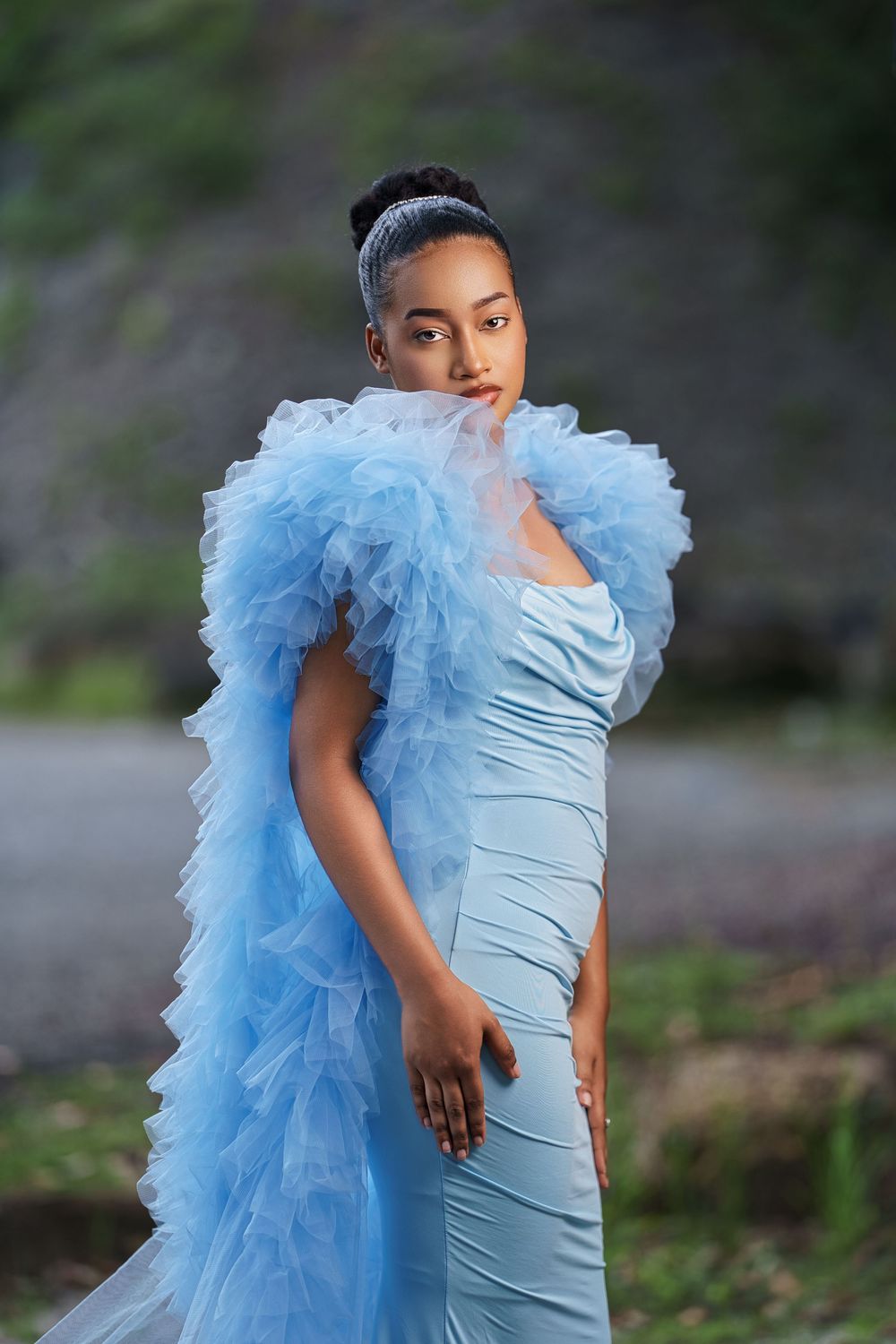 A model poses in a light blue gown with dramatic ruffled tulle detail against a blurred outdoor background.