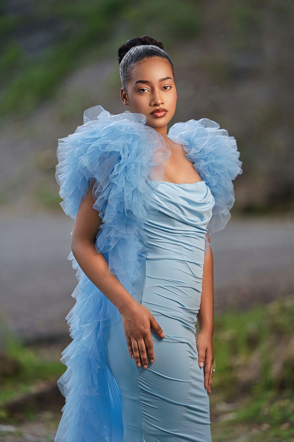 A model poses in a light blue fitted dress with dramatic ruffled tulle detail against a natural outdoor background.