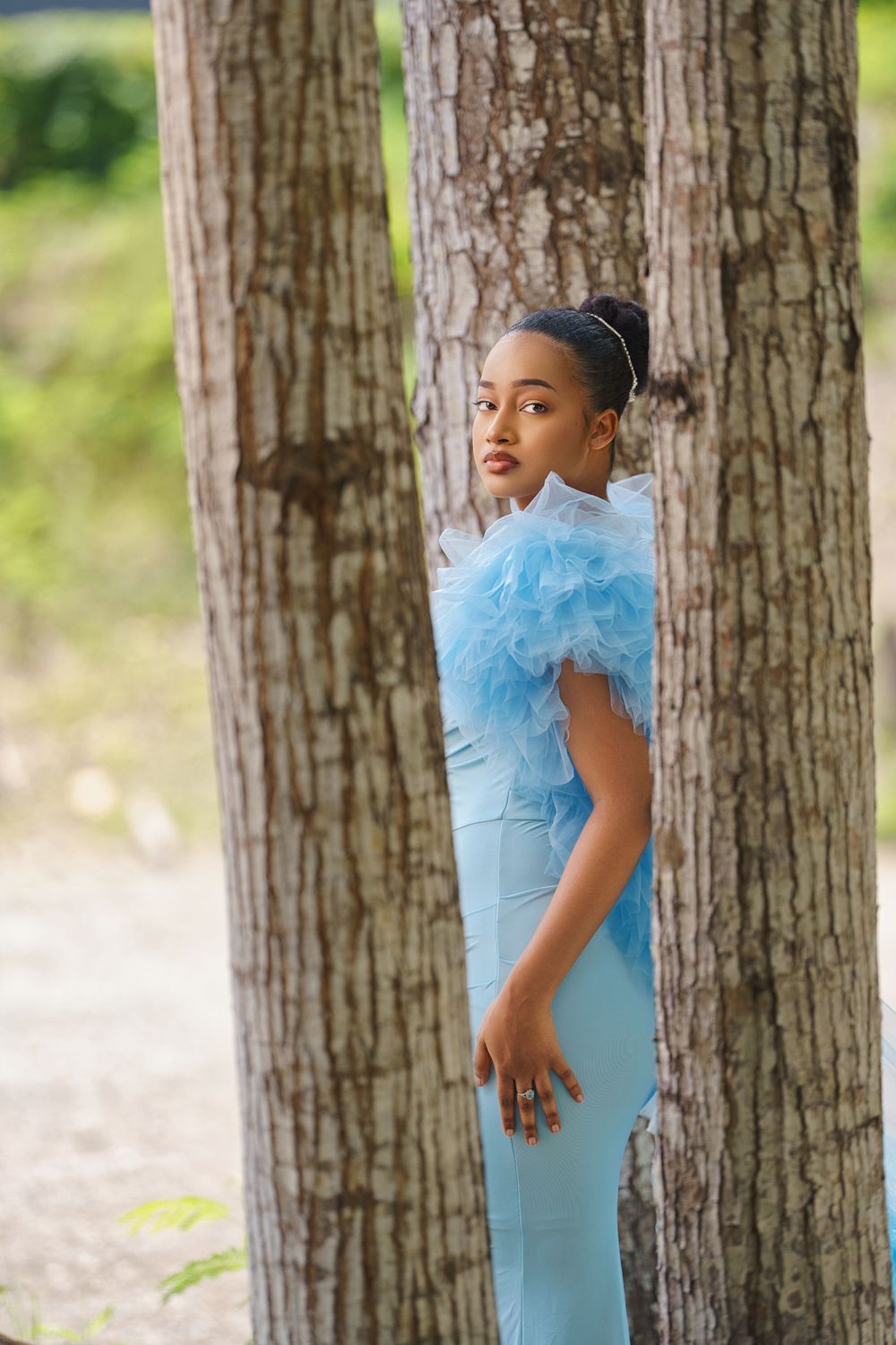 A child in a light blue dress plays hide and seek behind tree trunks in a wooded outdoor setting.