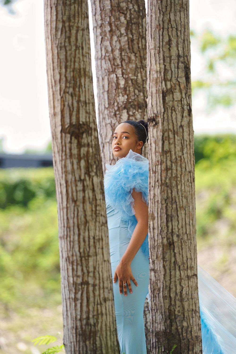 A figure in a flowing light blue dress poses thoughtfully between tall tree trunks in an outdoor setting.