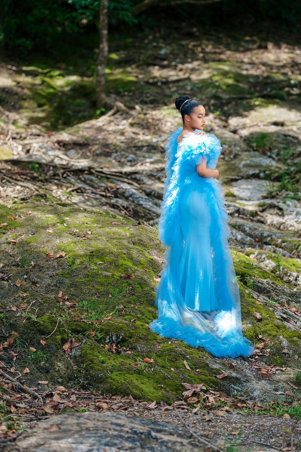 A figure in a flowing powder blue princess gown walks gracefully through a mossy woodland setting.