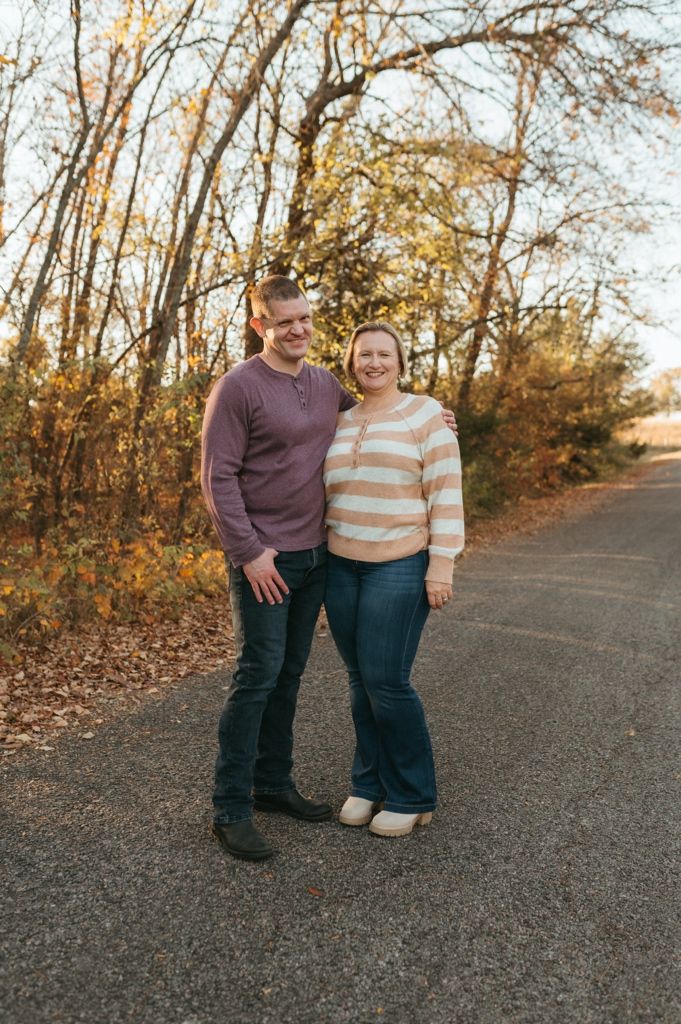 A couple stands together on a paved path surrounded by autumn trees during a golden hour photoshoot.