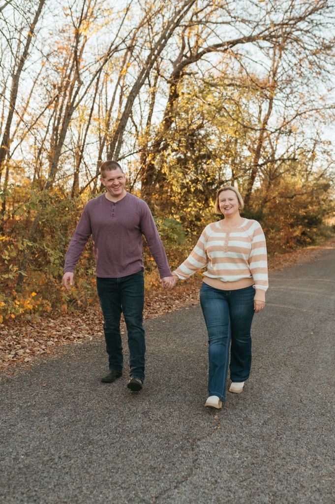 A couple walks hand in hand along a wooded path during golden hour in autumn.