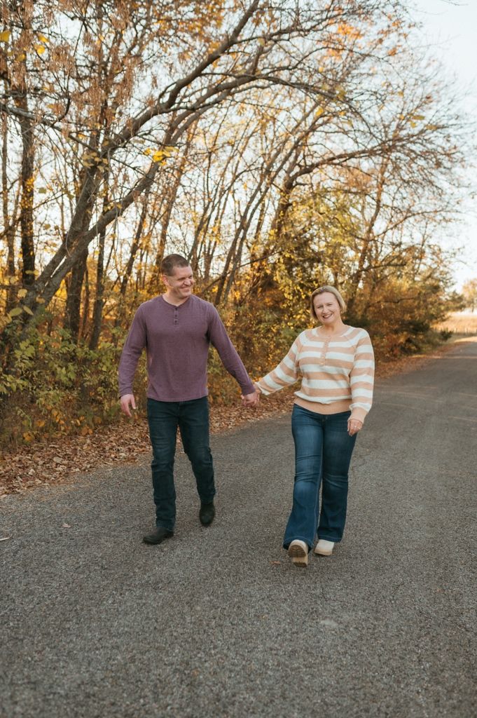 Two people walk hand in hand down a gravel path surrounded by autumn trees in warm golden light.