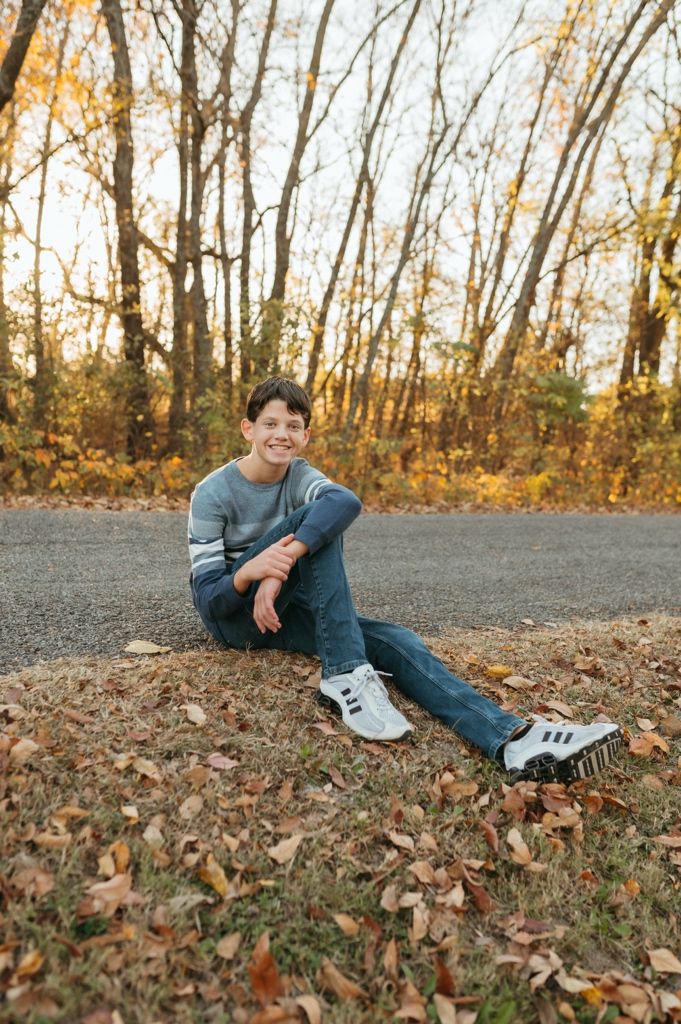 Young person lying on fallen autumn leaves on a path surrounded by trees in warm fall lighting.