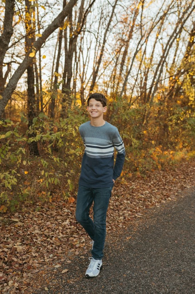 A child walks along a wooded trail in autumn, wearing a blue striped sweater and jeans among fallen leaves.