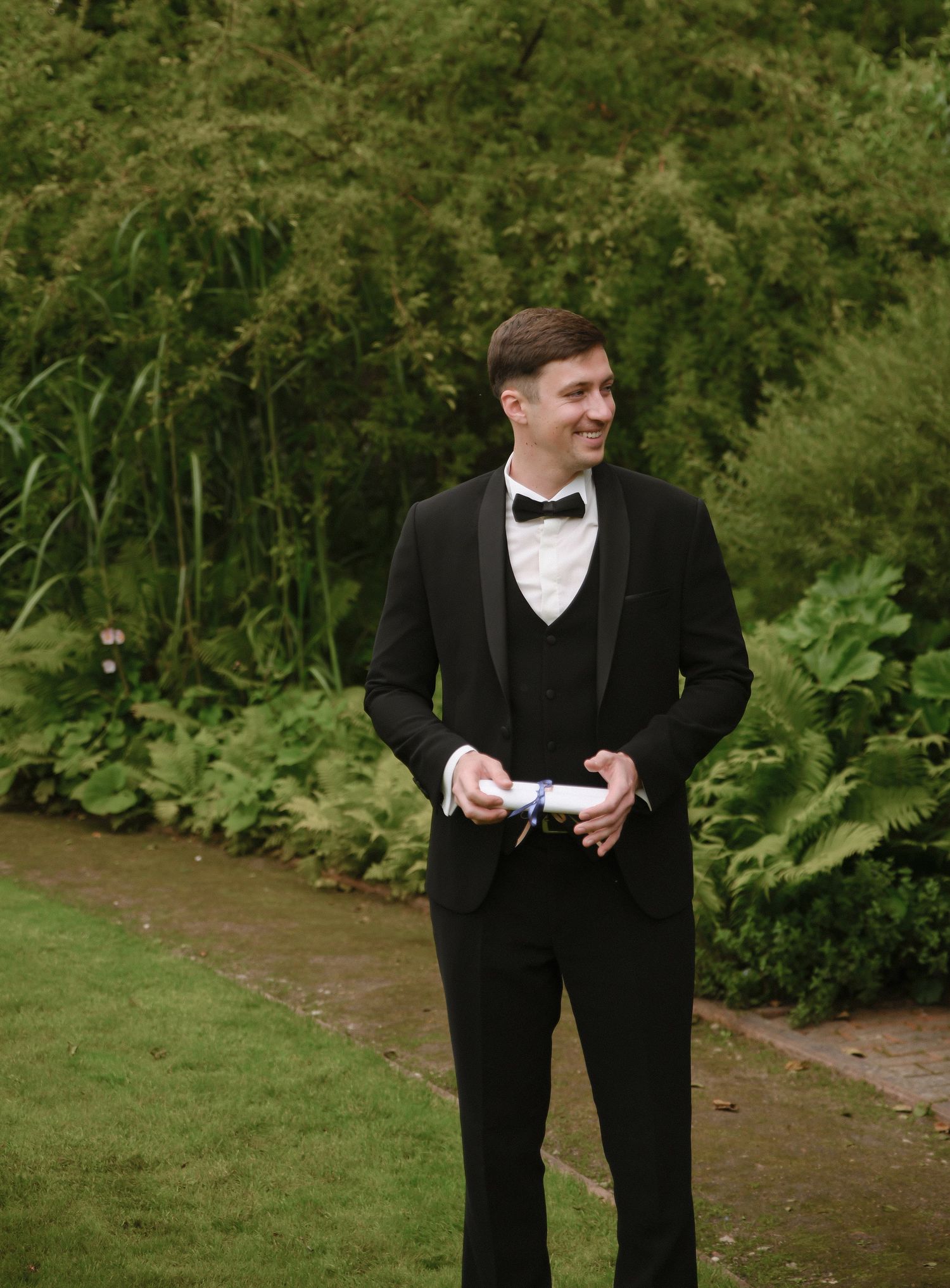 A person in a classic black tuxedo and bowtie stands on a garden path surrounded by lush greenery.