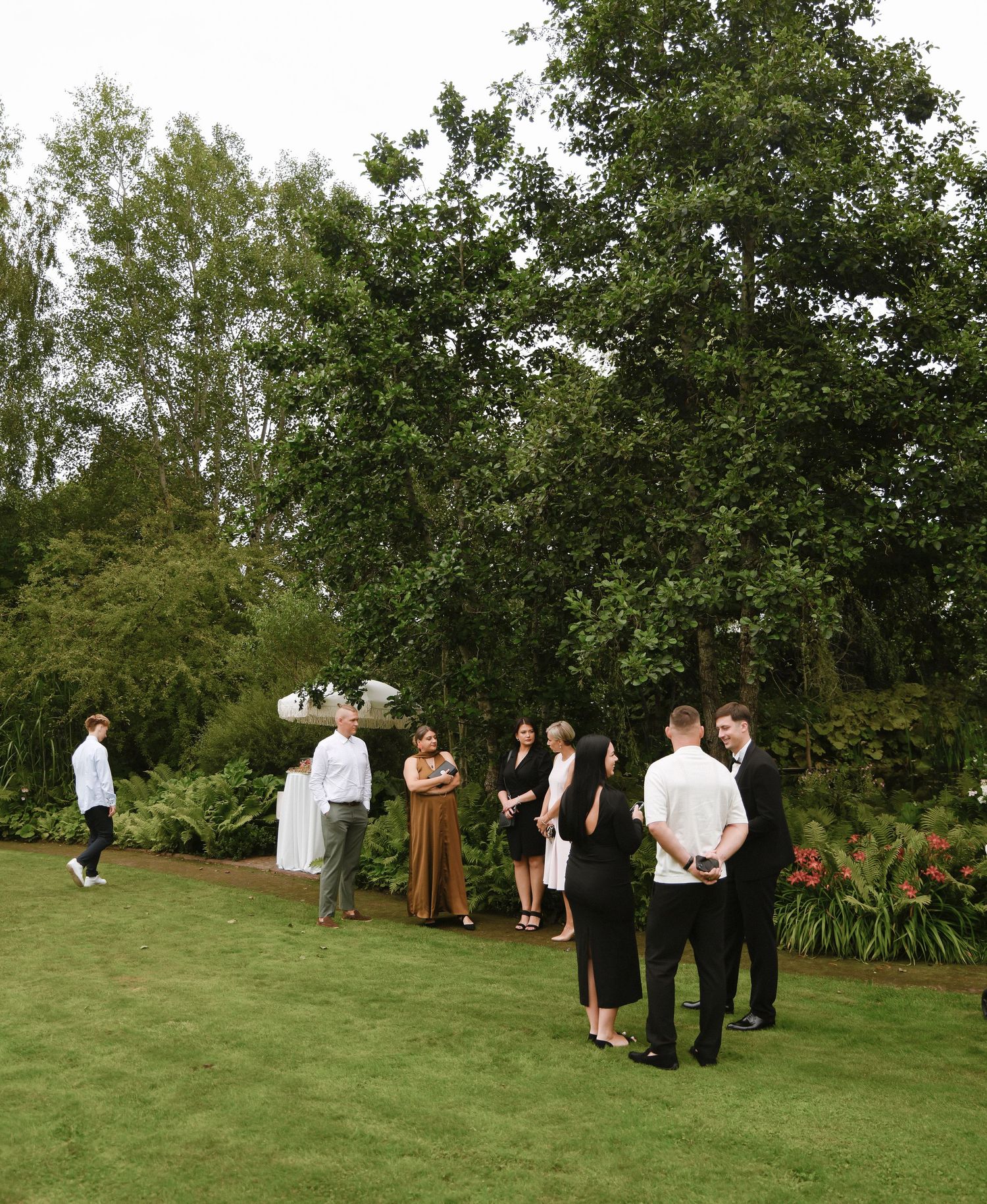 Wedding party members gather on a lush green lawn during an outdoor ceremony at a garden venue.