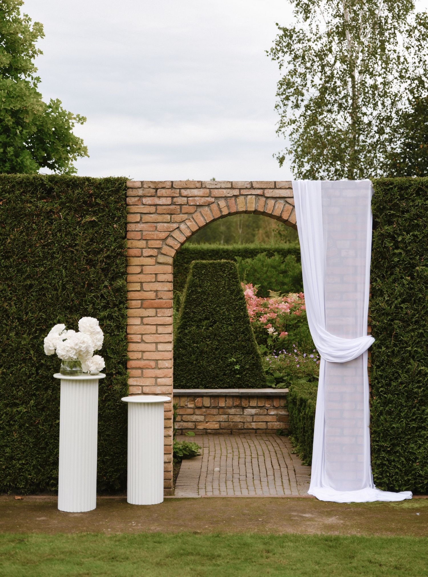 An elegant garden wedding arch with brick columns, white draping, and decorative urns beside a manicured hedge wall.