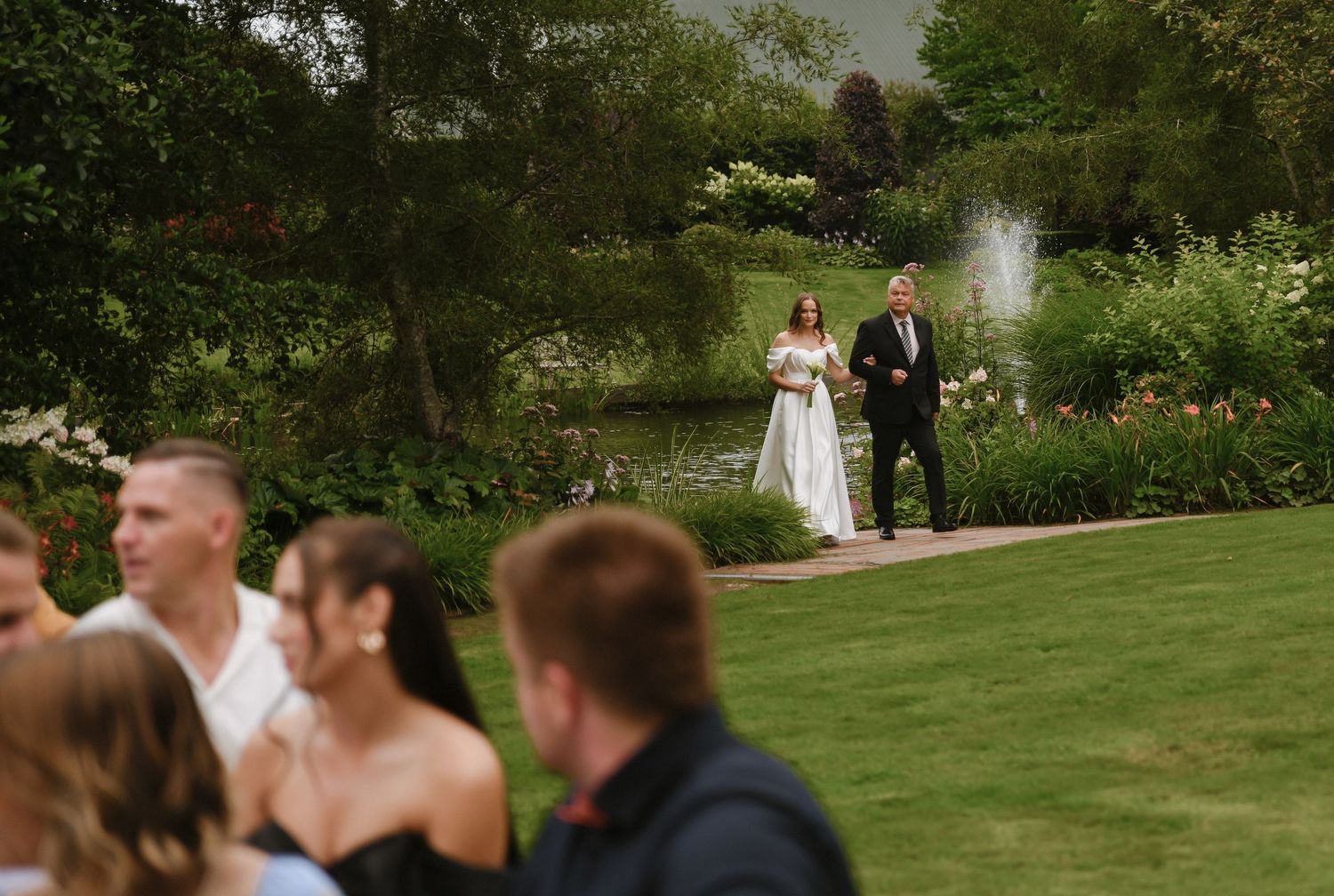 A wedding ceremony takes place on a lush green lawn beside a pond with trees and guests in attendance.