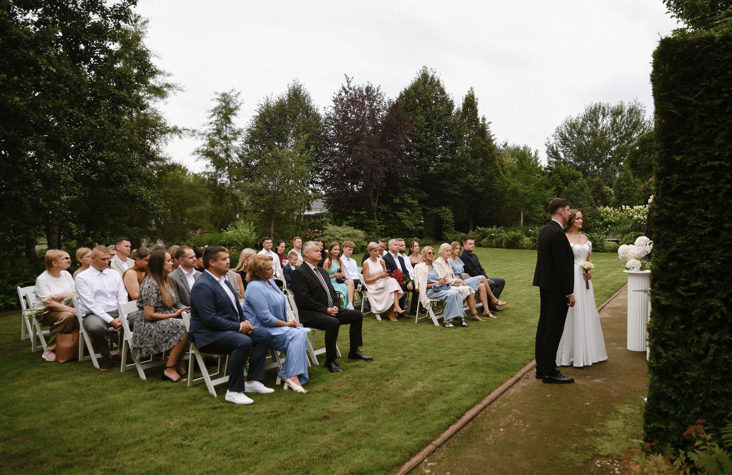 Outdoor wedding ceremony setup with rows of white chairs on a green lawn surrounded by trees.