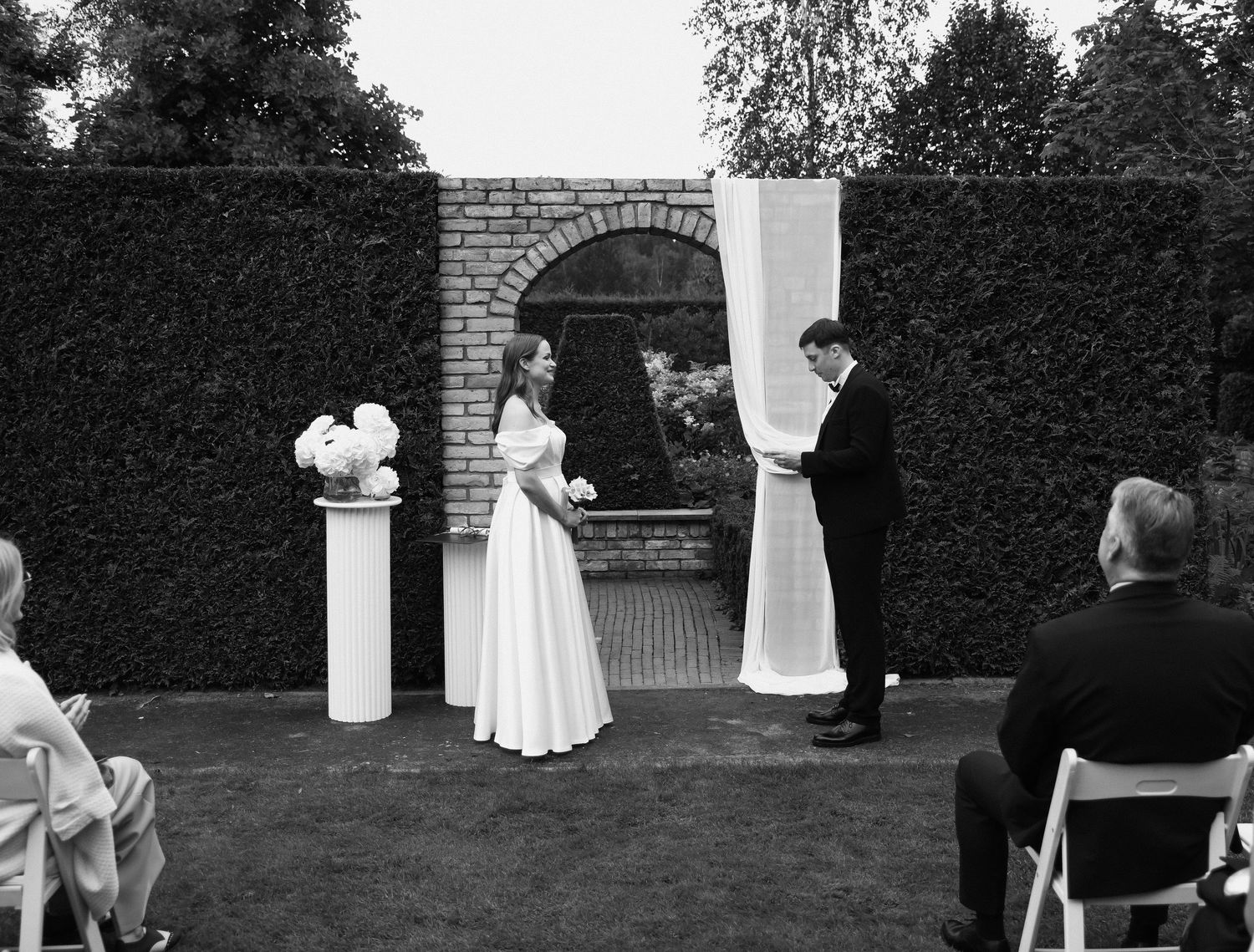 A black and white wedding ceremony scene with guests and decorations in an outdoor garden setting.