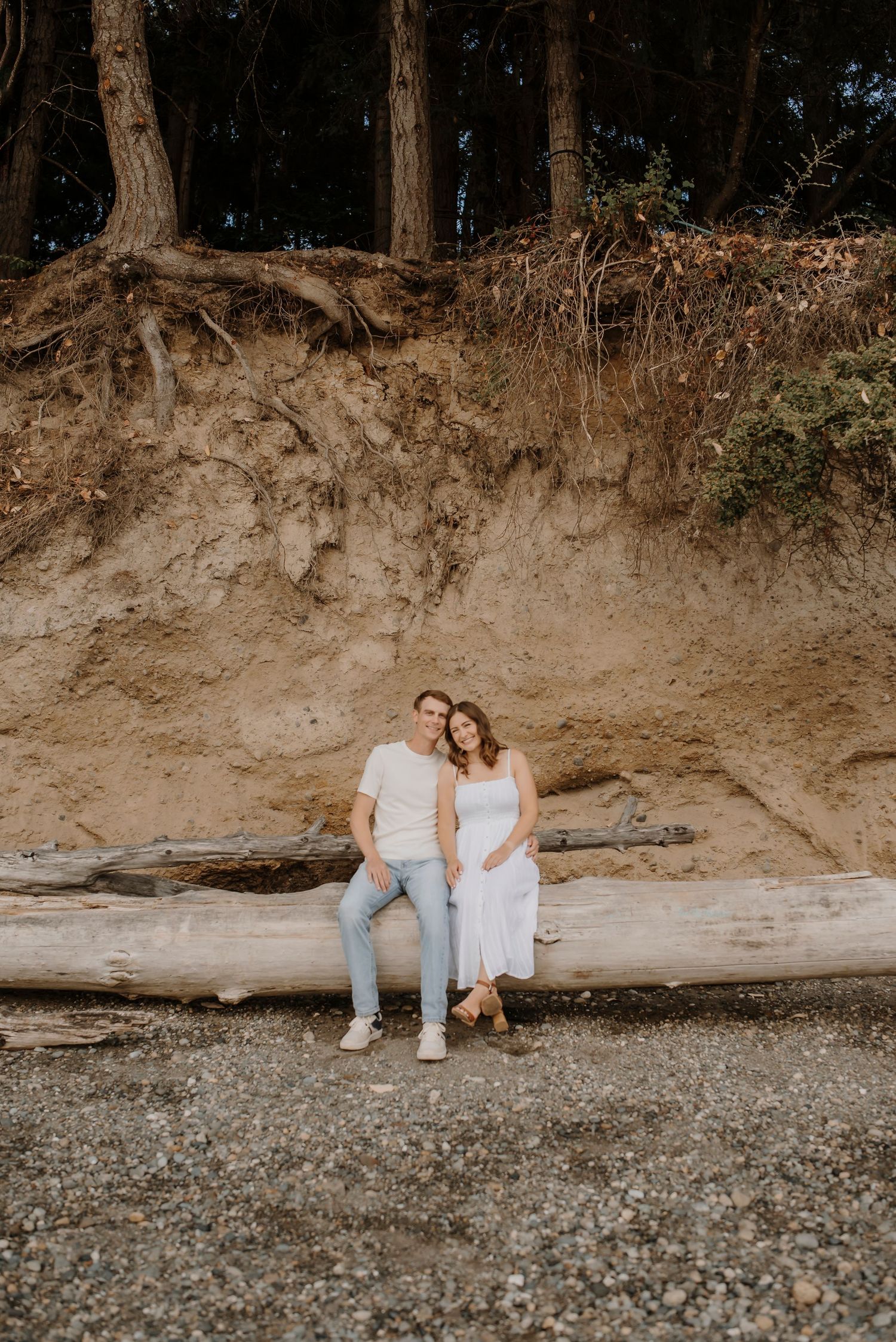 Couple sitting on rocky beach with forest backdrop during sunset.