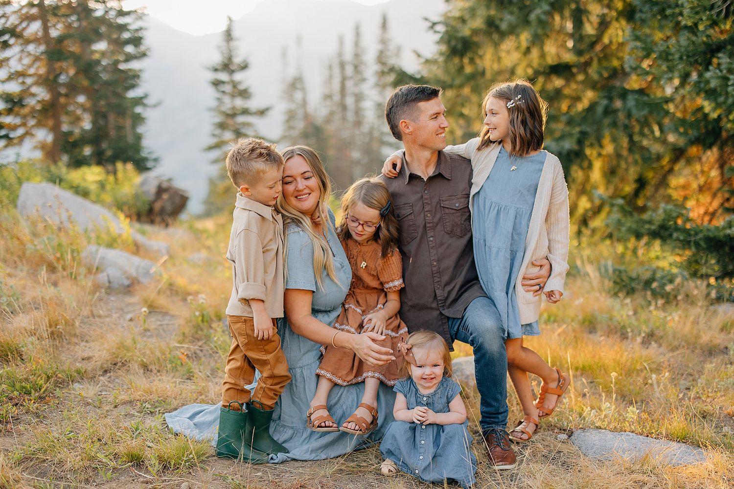 A family group poses together on a hillside with evergreen trees and golden autumn light in the background.