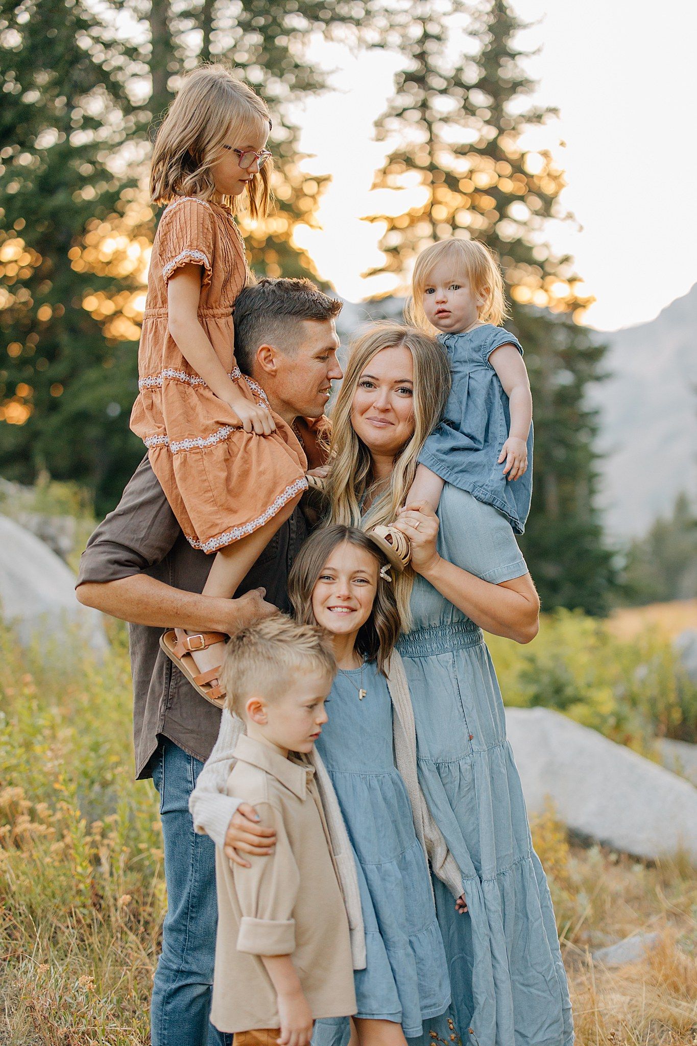 A family shares joyful moments together outdoors during golden hour sunset in a mountain setting with evergreen trees.