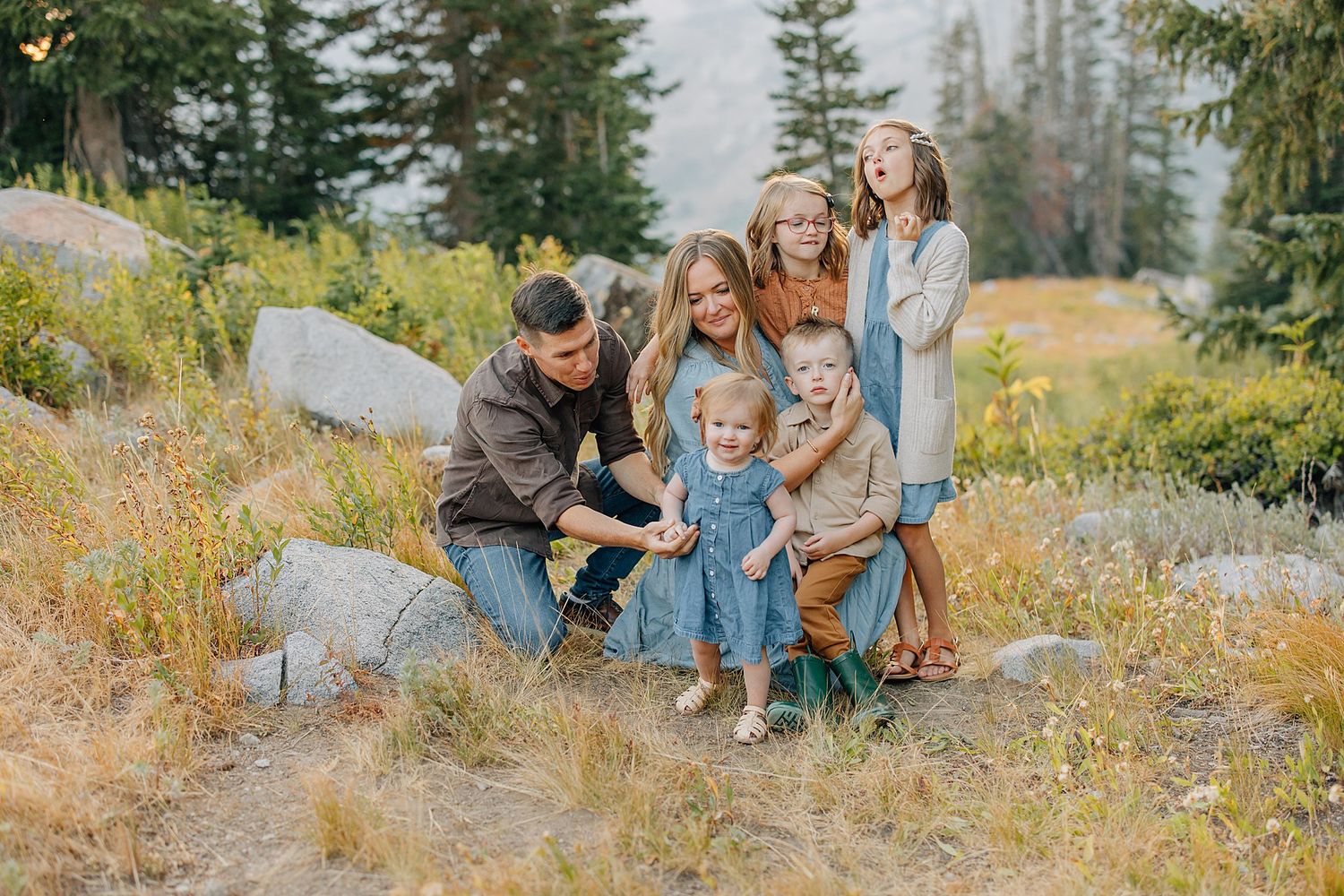 Family sits together on a grassy hillside surrounded by evergreen trees and autumn wildflowers on a sunny day.