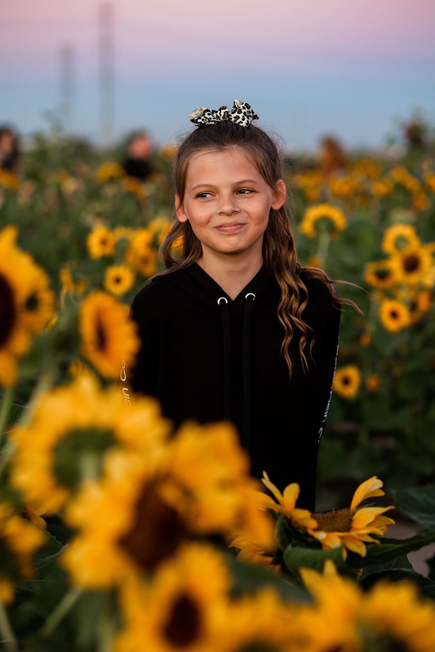 Smiling young girl with hair bow stands among vibrant yellow sunflowers at dusk.