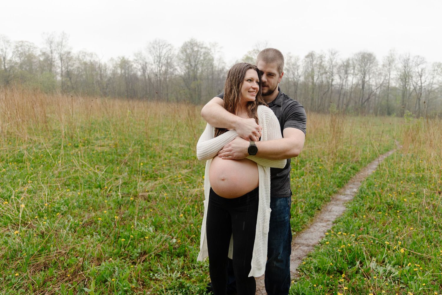 A couple embraces in a scenic field with trees in the background during an outdoor photo session.