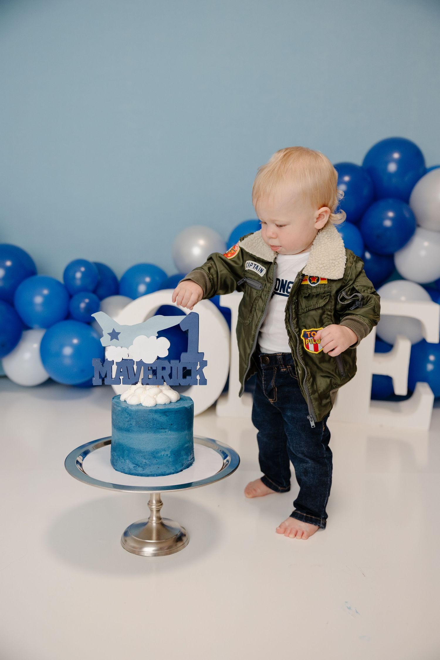 Baby in olive green jacket and jeans explores blue and white cake during first birthday cake smash photo session.