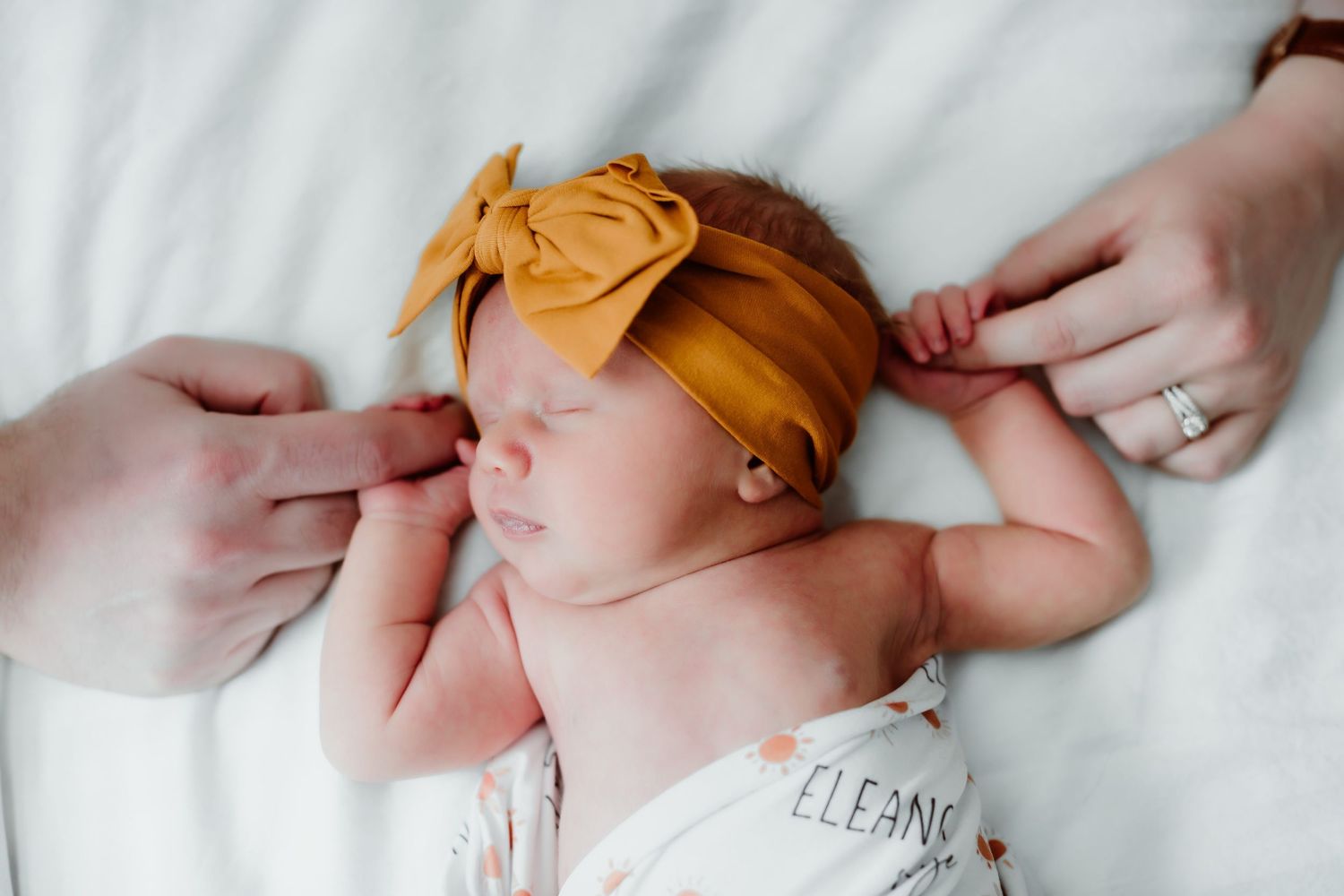 A newborn baby wearing a mustard yellow headband rests peacefully on white bedding.