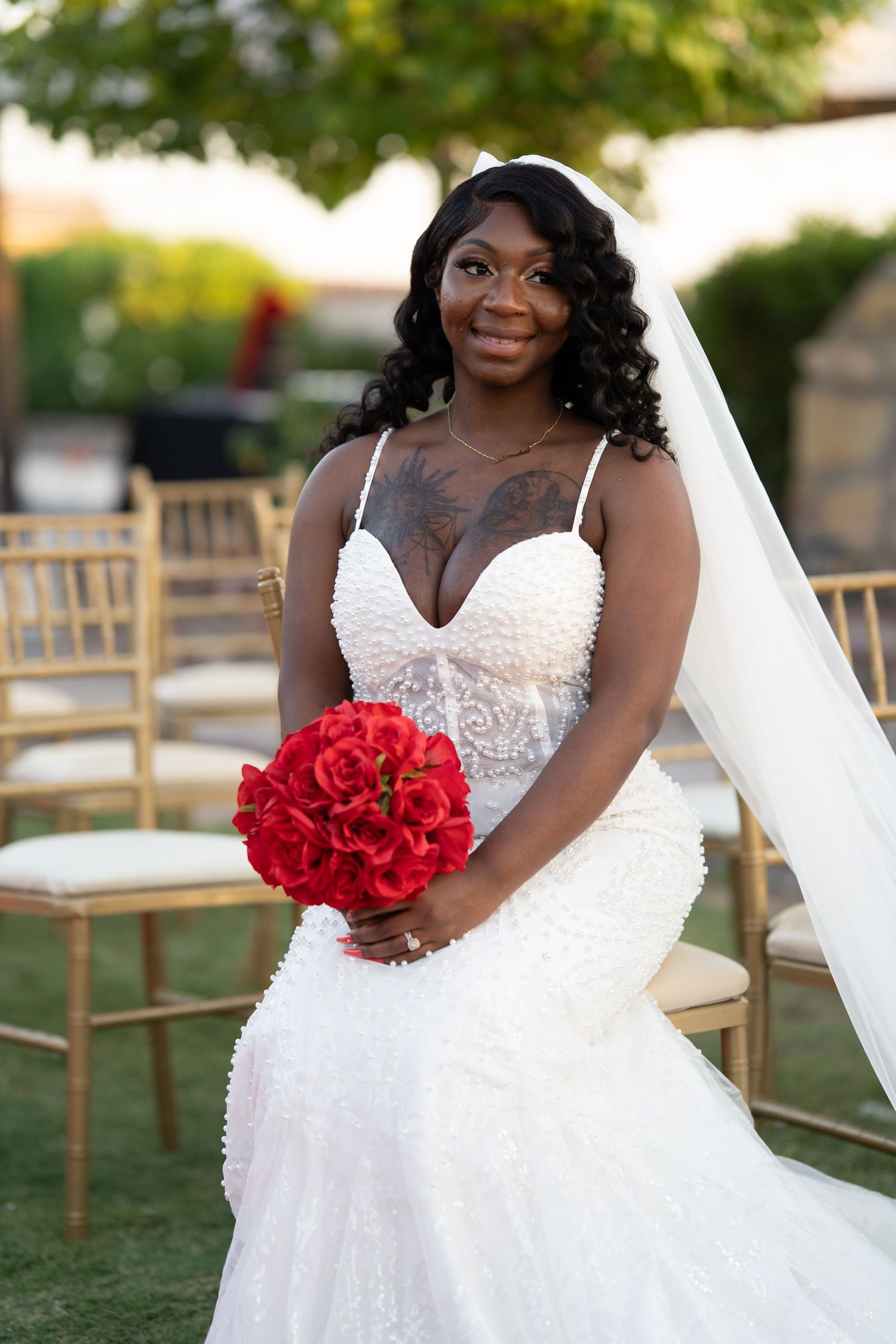 Bride in elegant white wedding dress poses with red rose bouquet against outdoor garden backdrop.