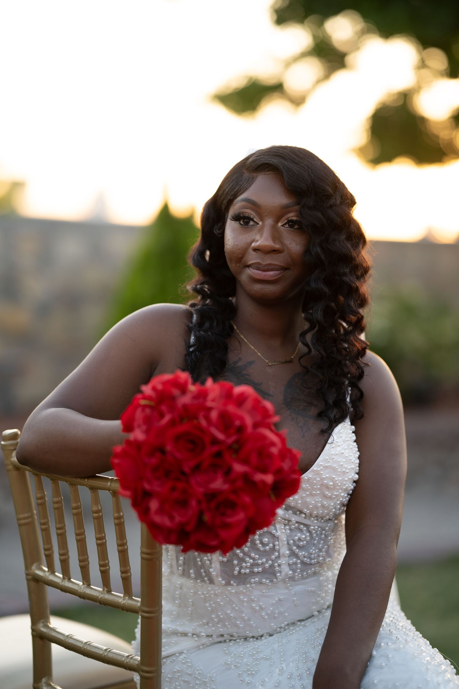 Beautiful bride in white lace wedding dress holds vibrant red rose bouquet while seated at sunset.