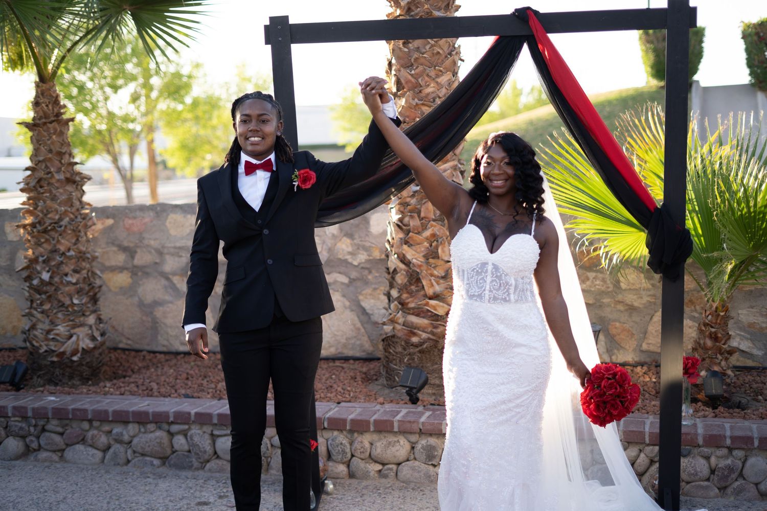 A wedding couple poses together under palm trees at an outdoor venue with red and black accents in their attire.