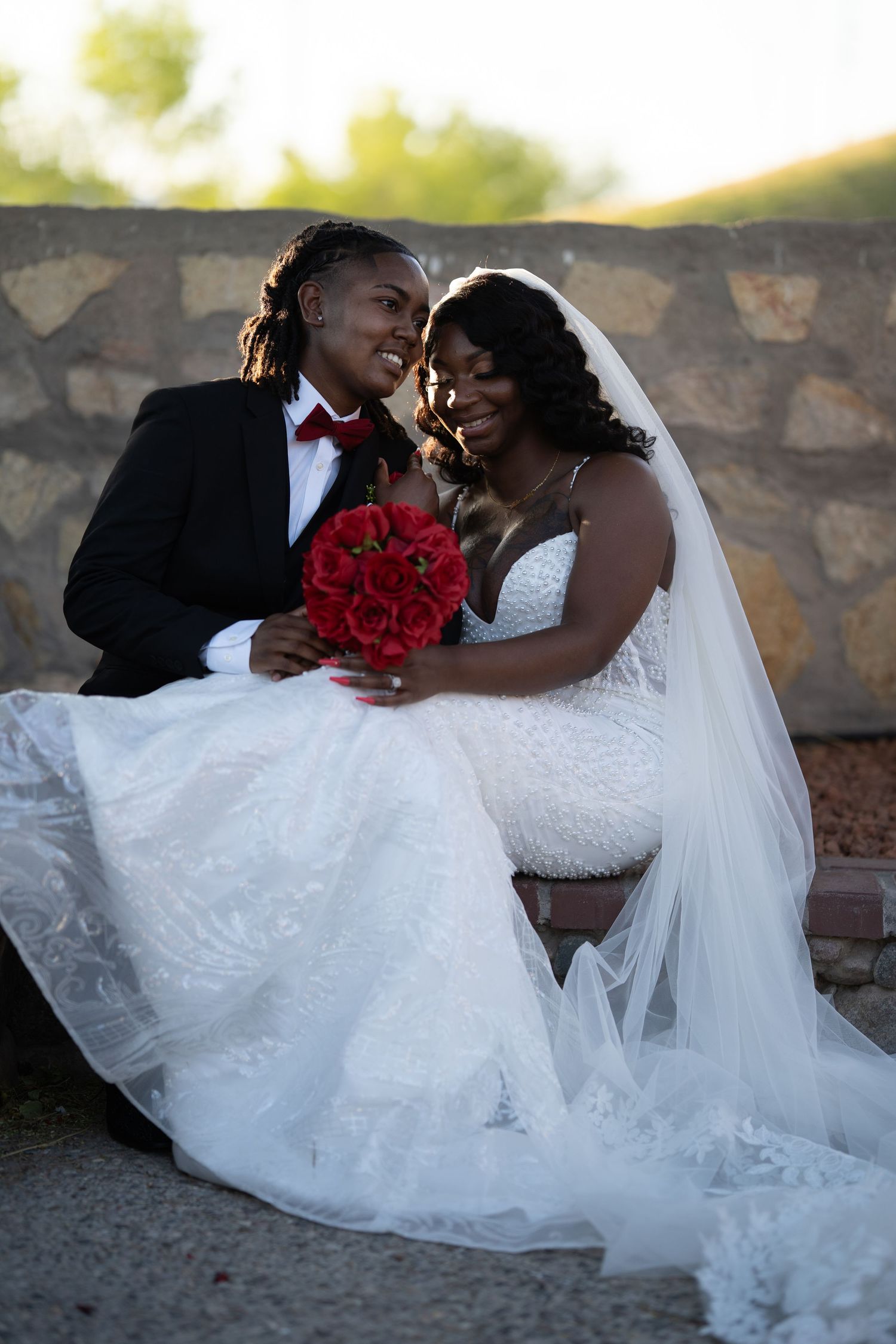 A romantic wedding couple in formal attire share intimate moments while posing with red roses against a stone wall.