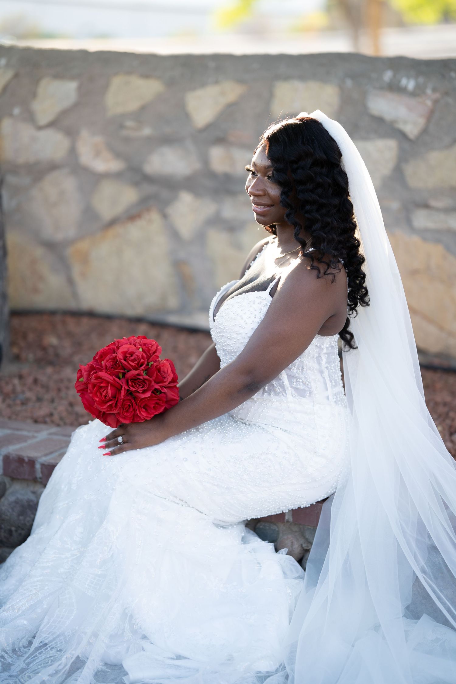 A bride in a white wedding gown with a flowing veil poses gracefully against a stone wall background.