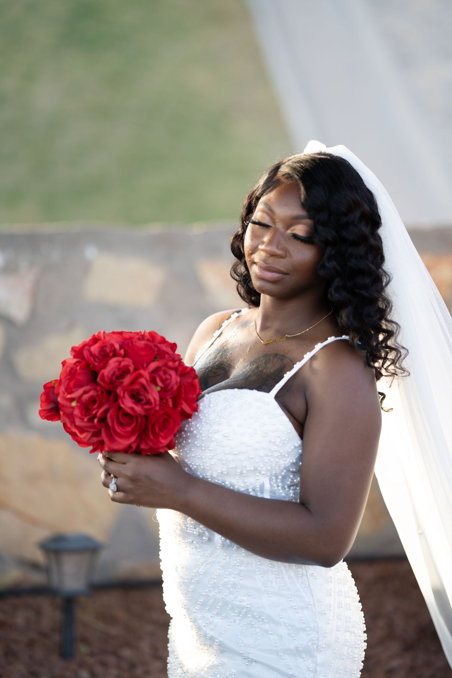 A bride in a white wedding dress holds a vibrant red rose bouquet while smiling with her eyes closed.