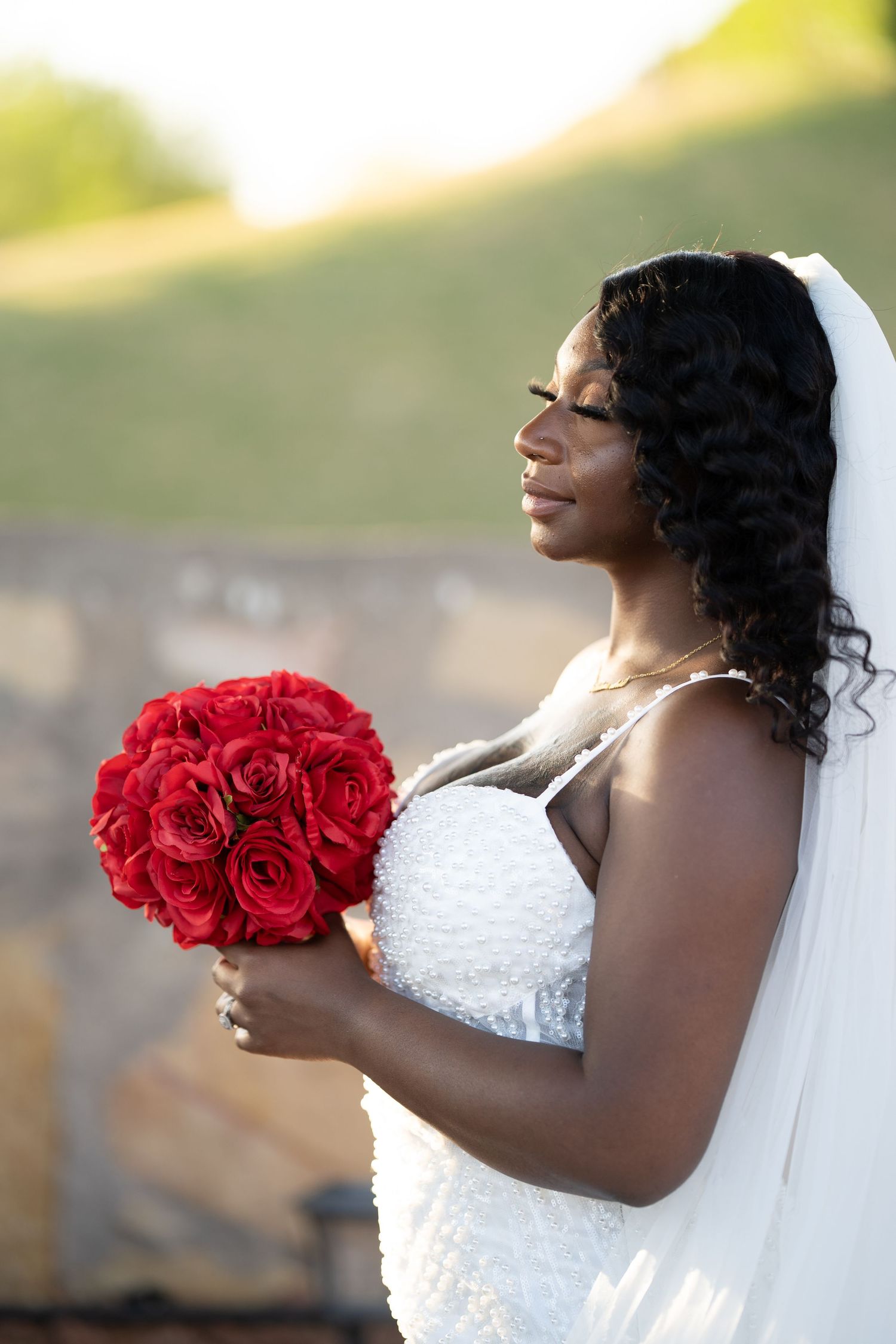 A bride in a white gown holds a bright red rose bouquet while looking to the side with a joyful expression.