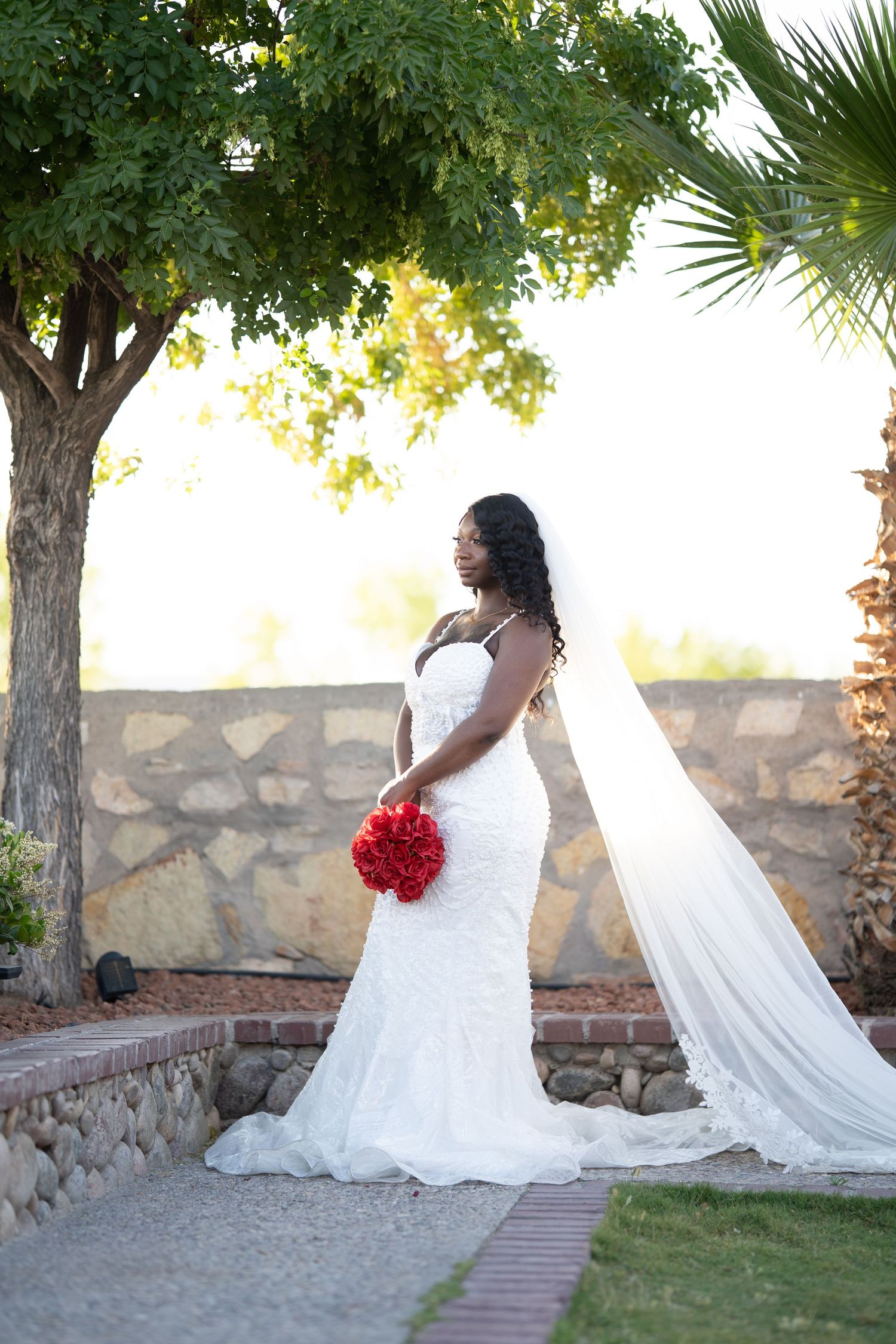 Elegant white wedding dress with flowing veil captured in a garden setting with stone walls and palm trees.