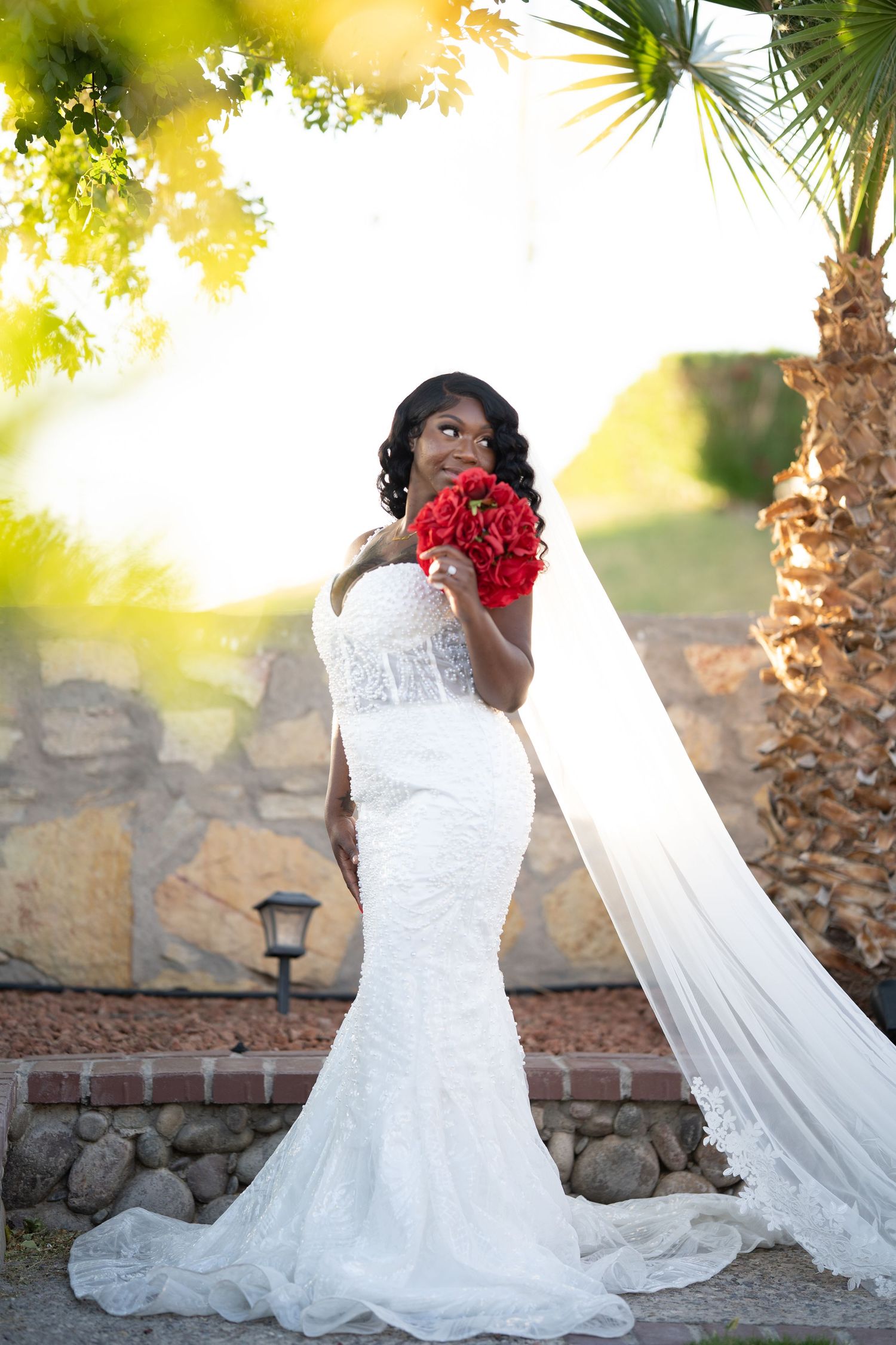A bride in a stunning white lace mermaid gown holds red roses against a palm tree and stone wall backdrop.