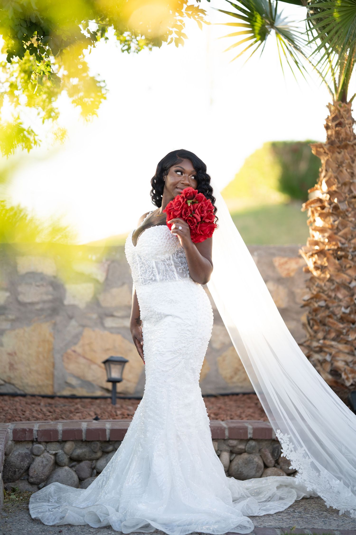 A bride in a stunning lace mermaid wedding gown holds vibrant red roses against a sunlit outdoor backdrop with palm trees.