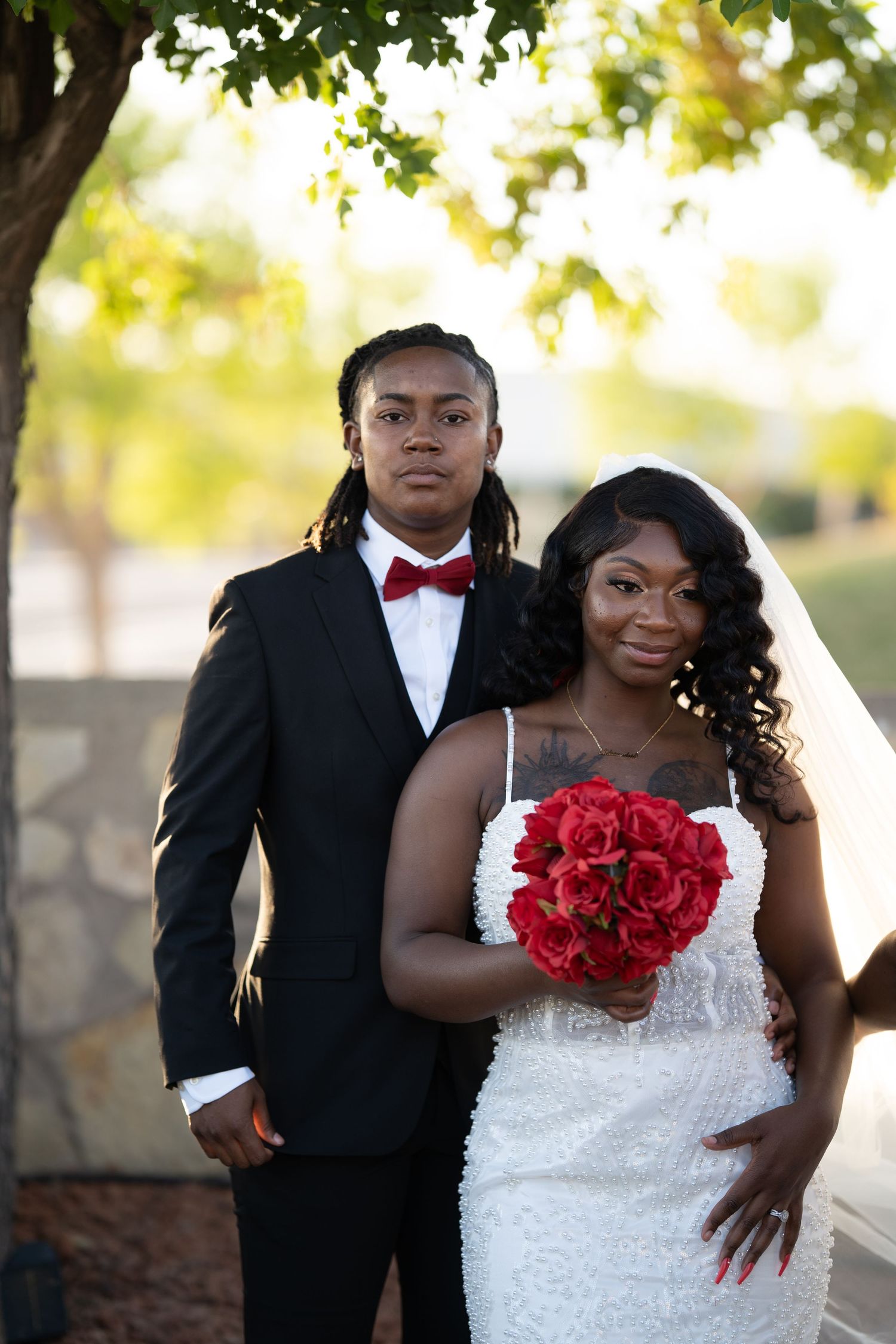 Wedding couple poses together outdoors with the bride wearing a white lace gown and holding vibrant red roses.