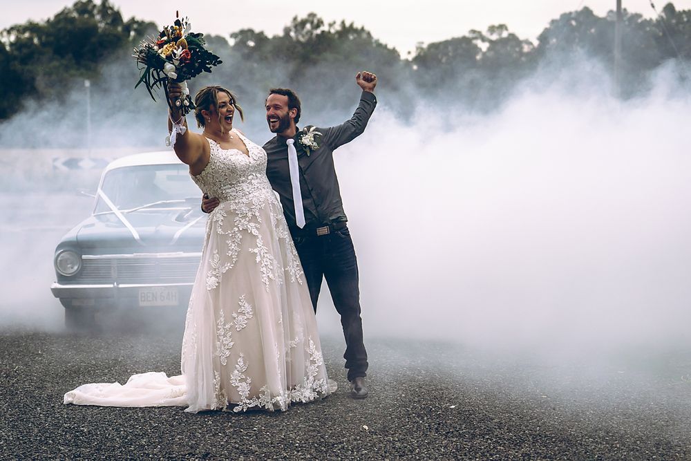 A couple celebrates their wedding day with joyful poses in a misty outdoor setting with a vintage car in the background.