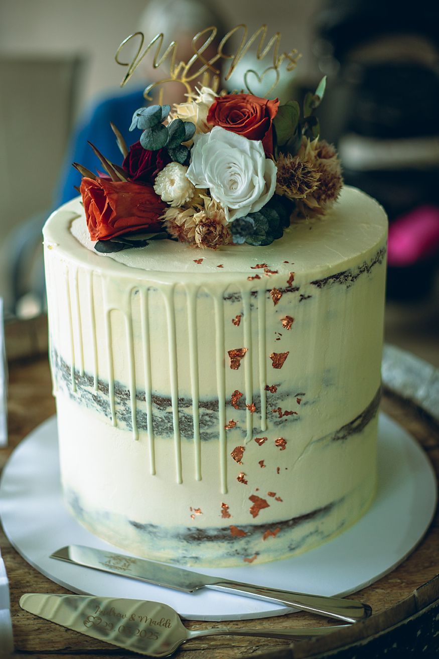 A white wedding cake with dripping icing decorated with fresh red and white flowers on top.