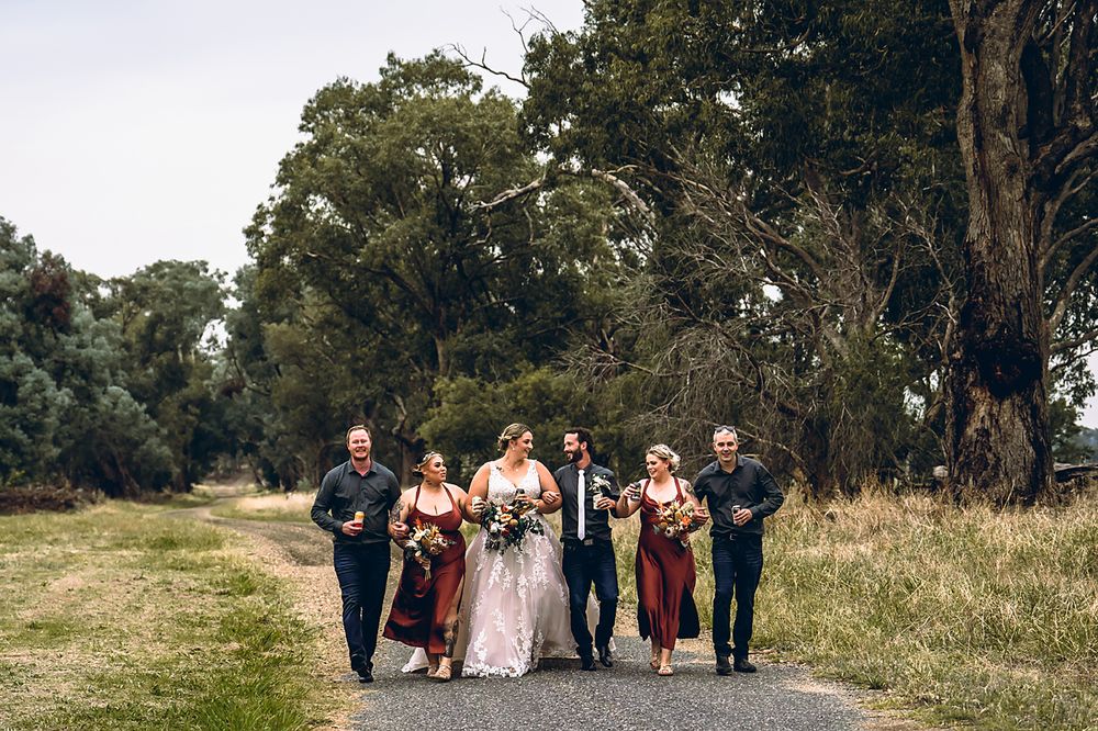 Wedding party walks together in a line through a scenic outdoor field with tall trees in the background.