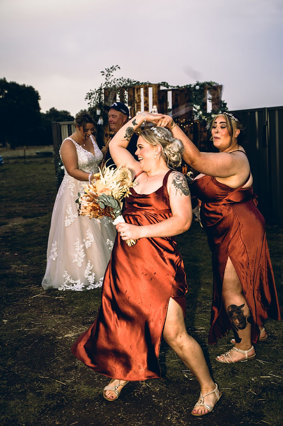 Bridesmaids in rust-colored dresses playfully dance and celebrate together in an outdoor setting at sunset.