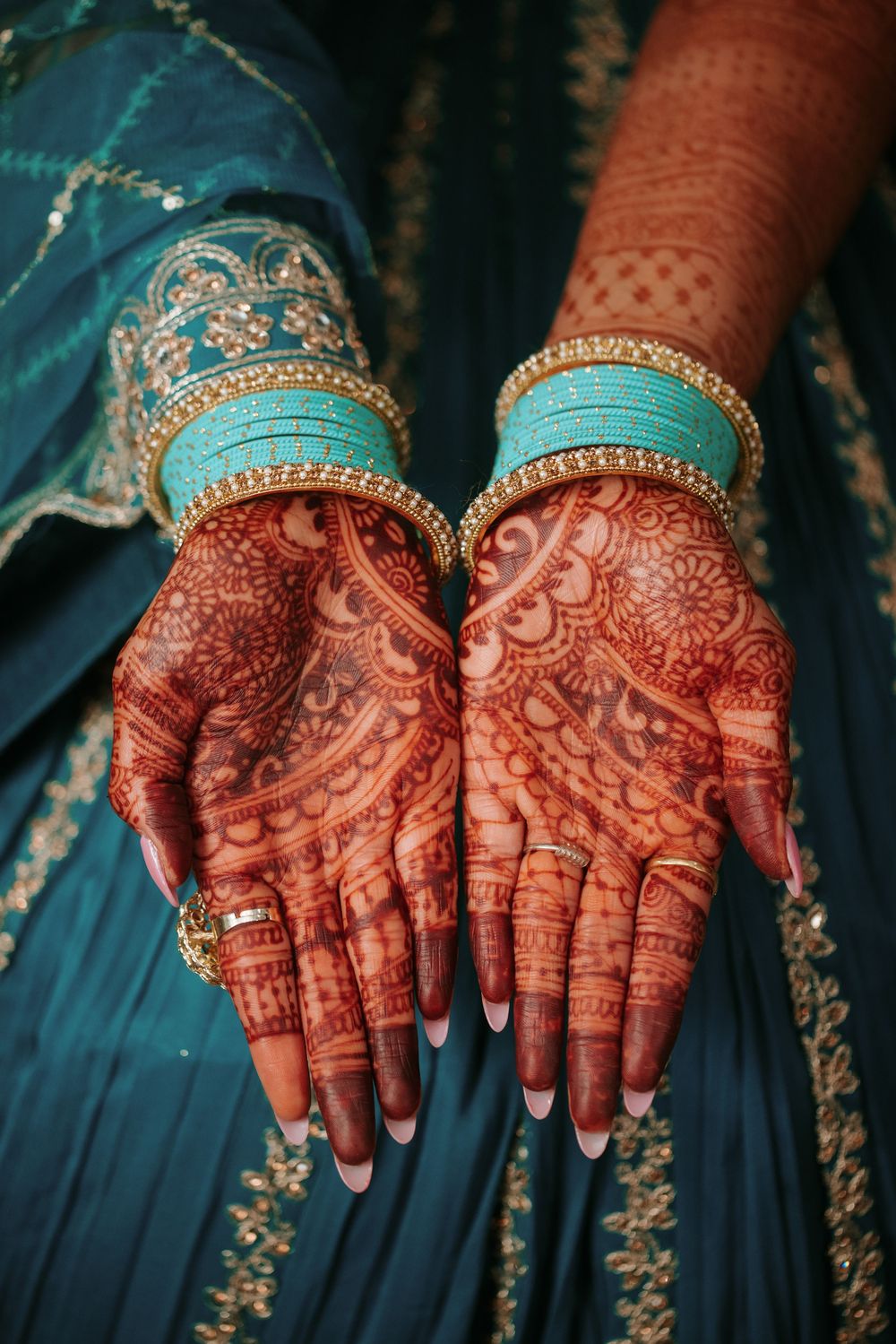 Intricate henna designs adorn hands wearing traditional Indian bridal bangles against a turquoise background.