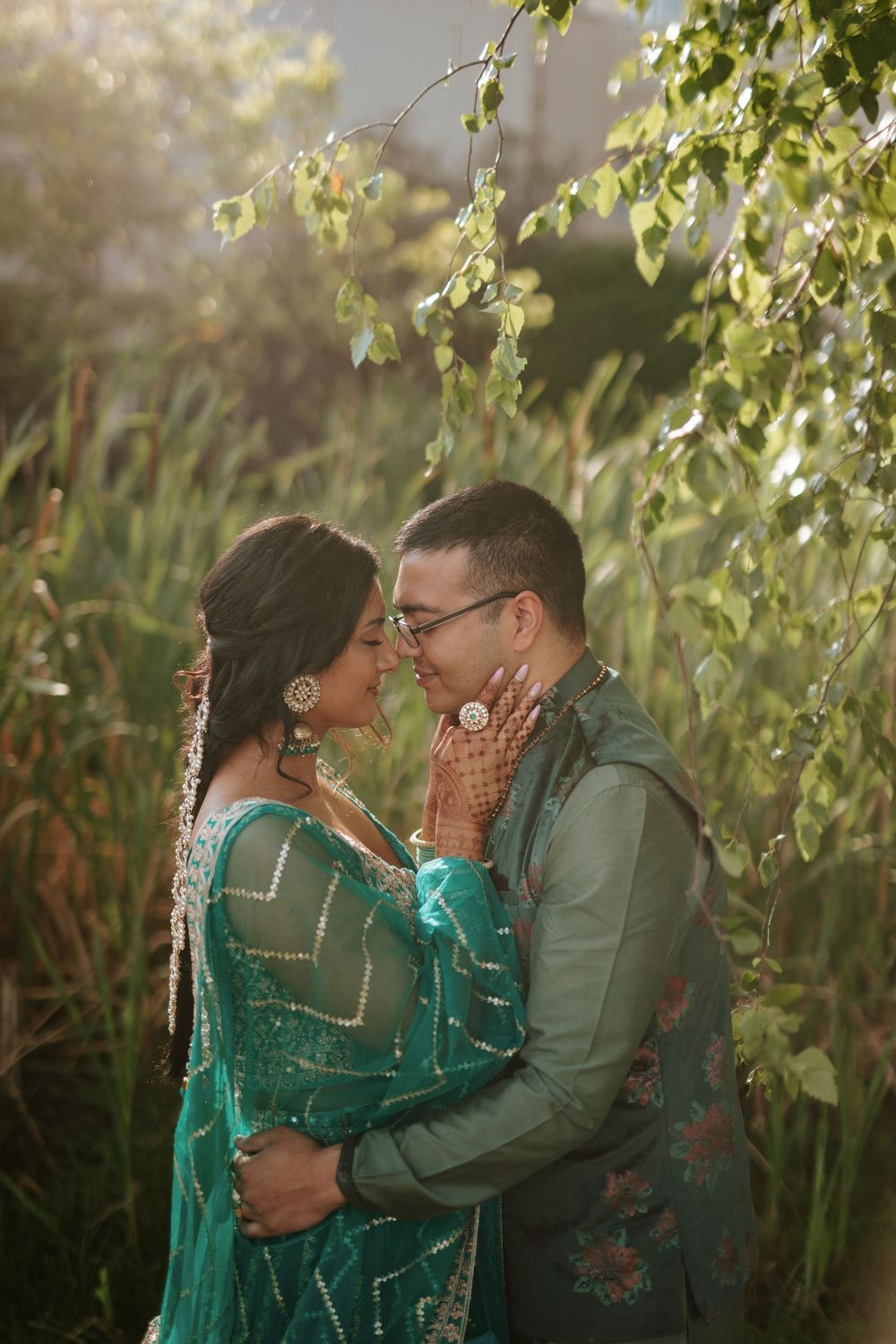Romantic couple in emerald green attire embracing outdoors among sunlit foliage.