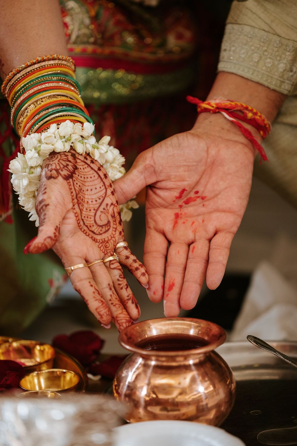 Close-up of hands decorated with traditional henna and bangles performing rituals with copper vessels in an Indian ceremony.