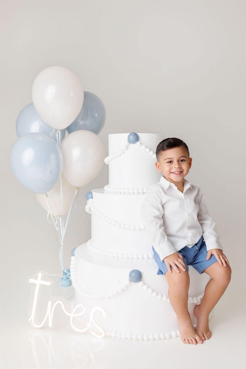 A cake smash photo session setup with white cake, blue and white balloons against a light gray backdrop.