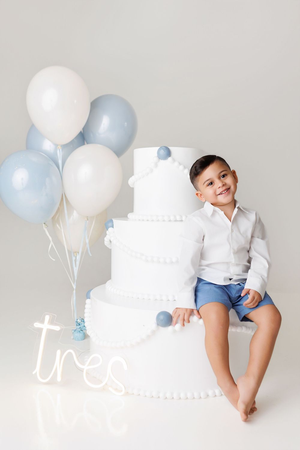 Cake smash photo session with blue and white balloons featuring a white tiered cake against a light gray backdrop.