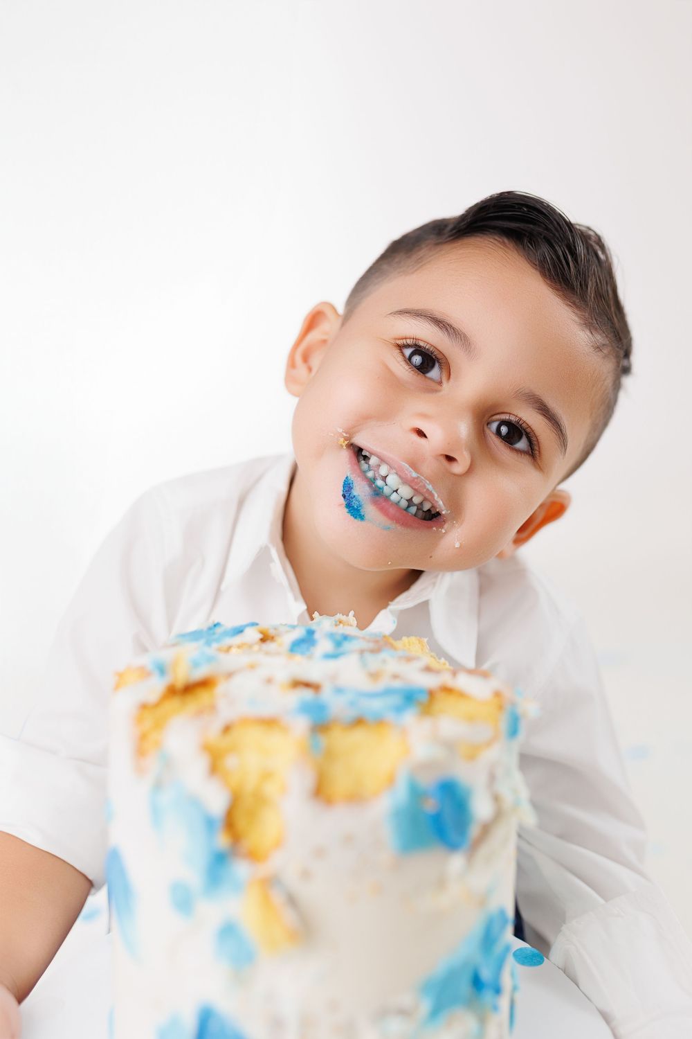 Playful young person in white shirt with blue braces and frosting on face smiling next to decorated cake.