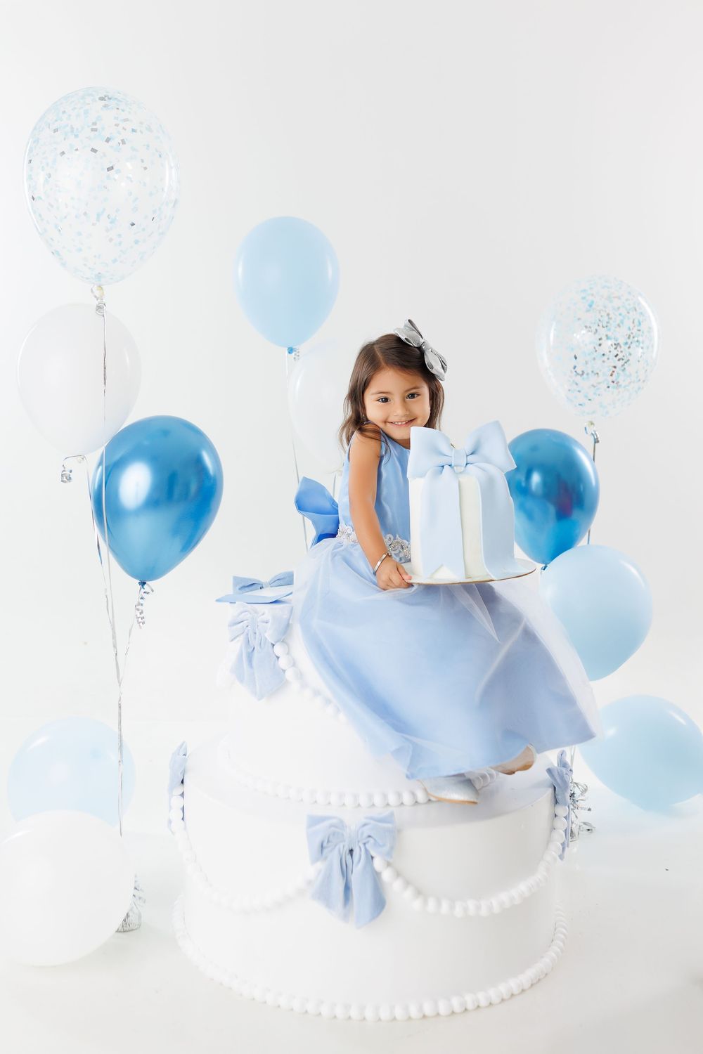 A child in a blue dress sits on a large white cake prop surrounded by blue and white balloons for a photo session.