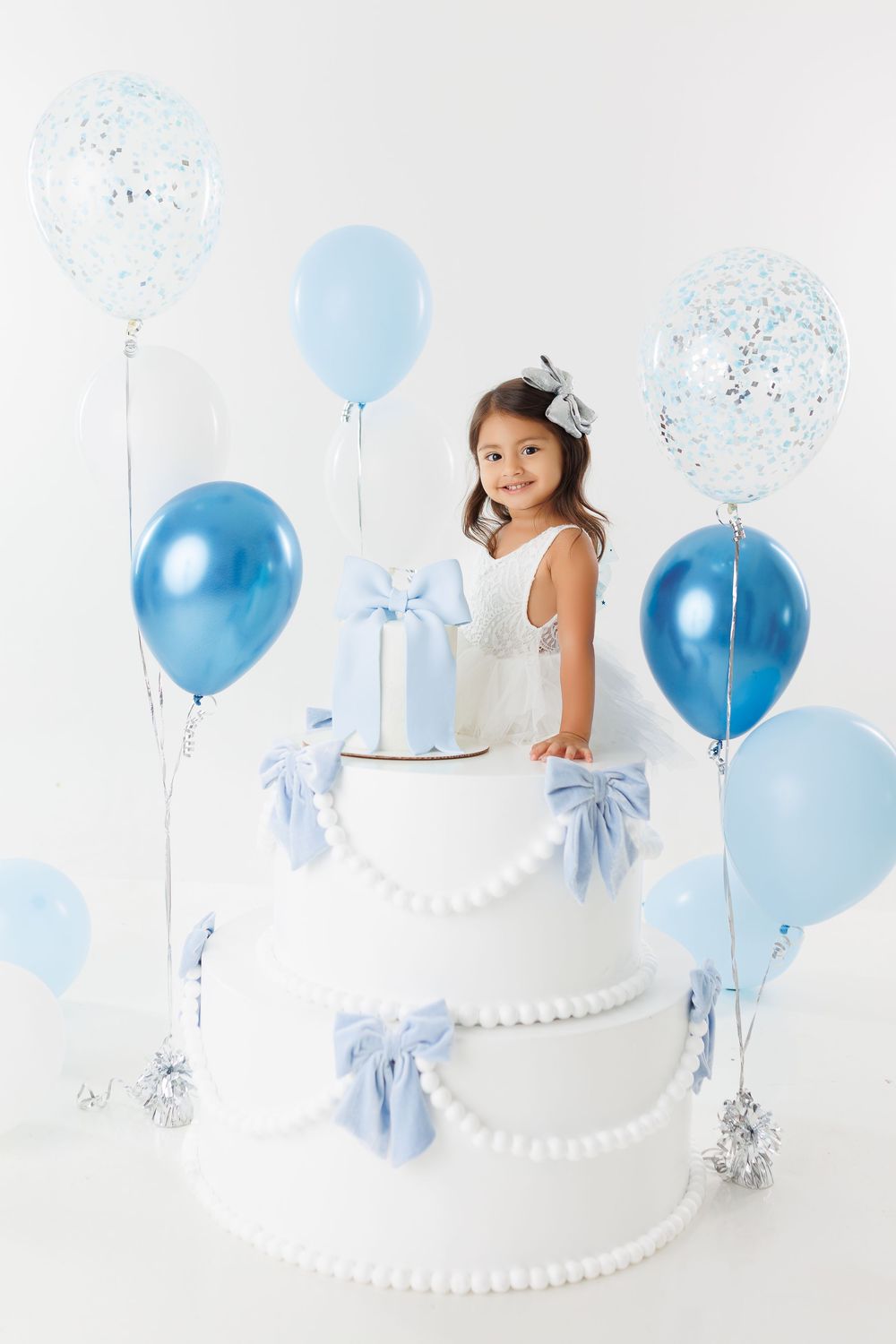 White tiered birthday cake with blue balloons and silver confetti decorations against white background.